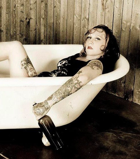 rachel, a tattooed white woman in a pin up style photoshoot where she lounges in a bathtub, arm held out holding a bottle. Her hair swirled around her face like betty boop.